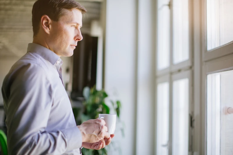 Mann steht mit Becher vor dem Fenster und guckt nach draußen